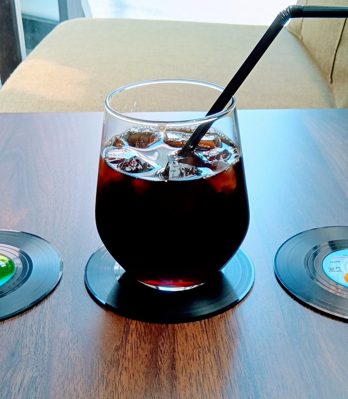 ダークウッドのテーブルにレコード盤型コースターを敷いて置かれた、氷入りアイスコーヒーのグラスと黒いストロー
